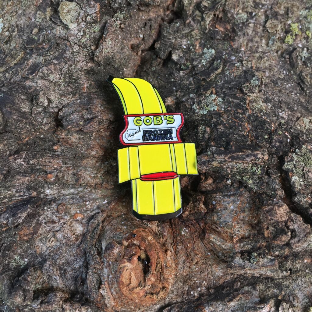 lapel pin in the shape of a banana stand that says "Gob's original frozen banana" sitting in a tree's knot