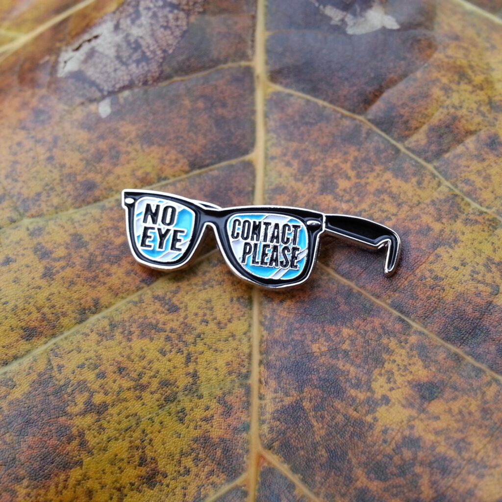 lapel pin of sunglasses that say "no eye contact please" on the lenses. stuck into a fallen fall leaf